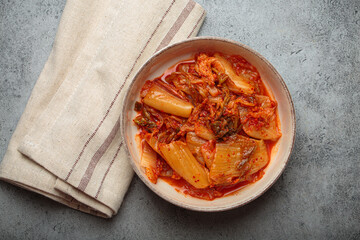 Kimchi, a fermented napa cabbage Korean dish, in a rustic ceramic bowl on a grey concrete background with a linen napkin, capturing its vibrant color and texture from a top down view