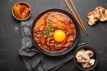 Top view of spicy kimchi jjigae soup in a black bowl with egg yolk, enoki, shiitake, tofu and green onions, with chopsticks and cooking ingredients, cuisine of Korea