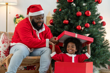 Happy boy with dad in Santa suit sitting inside big Christmas gift box. African American family celebrating Merry Christmas on holiday.