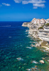 view of the medieval town of Bonifacio with its white limestone coastline and rough, deep blue turquoise sea and boat in sunshine and blue skies