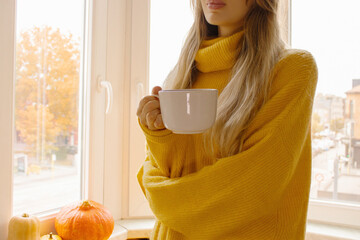 Woman in yellow knit sweater holding white mug by a window with pumpkins on the sill. Cozy autumn...