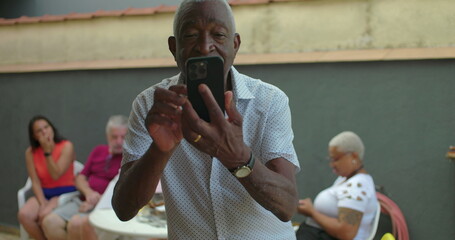 African American Elderly man smiling while looking at smartphone screen, standing outdoors with casual group conversation in blurred background, engaged with digital interaction