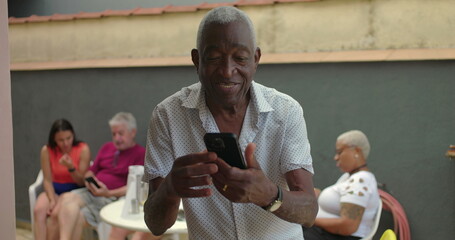One black Elderly man holding a smartphone outdoors, focused and thoughtful, interacting with the device, with diverse adults in the background at a social gathering