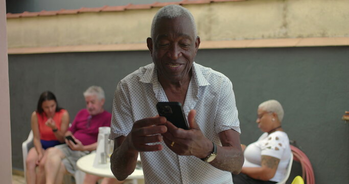 One black Elderly man holding a smartphone outdoors, focused and thoughtful, interacting with the device, with diverse adults in the background at a social gathering