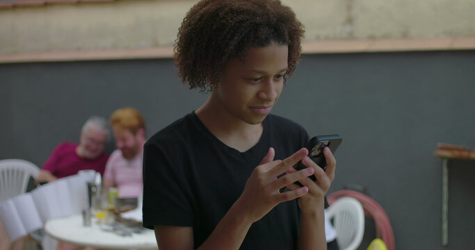 One young Teenager of African descent using a smartphone outdoors, focused and engaged, with diverse adults in the background enjoying a casual social gathering - Powered by Adobe