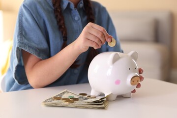 Little girl putting coin into piggy bank at white table indoors, closeup. Pocket money and responsibility