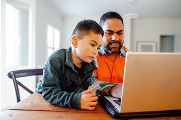 Father working on laptop on dining table while toddler son looks at screen beside him