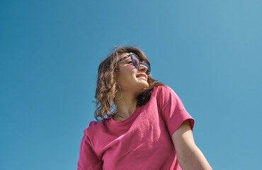 Beautiful carefree young woman in sunglasses and pink t-shirt smiling and looking aside on blue sky background low angle shot. Copy space