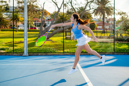 Young woman hits forehand shot on tennis court surrounded trees during daylight