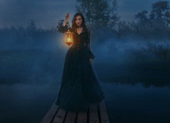 Fairy tale mystery woman queen hand hold lantern lamp candle walking across bridge pier over river dense fog mist dark blue night nature. Fantasy girl beauty face gothic makeup long hair black dress