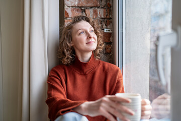 Serene woman relaxing takes coffee break looking to window with positive thoughts. Contented young female spending calm pause with pleasure, stress relieve, holding cup drink, resting in loft interior