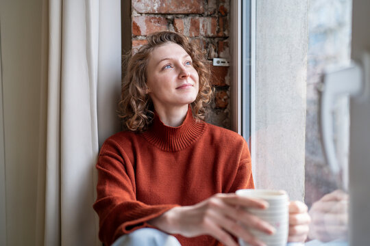 Serene woman relaxing takes coffee break looking to window with positive thoughts. Contented young female spending calm pause with pleasure, stress relieve, holding cup drink, resting in loft interior