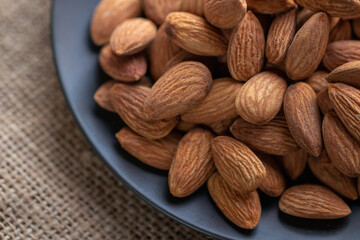 A closeup shot of a pile of raw almonds on a dark blue plate, with a burlap texture visible in the background
