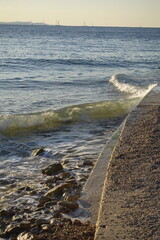 Coastal view with waves crashing against a stone pier and sailboats in the distance