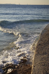 Waves crashing on a rocky shore with sailboats on the horizon at sunset