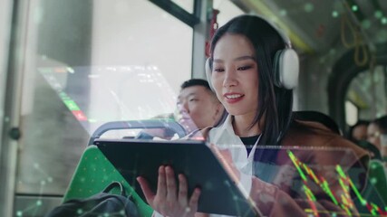 Asian woman wearing headphones sitting in bus. Holding tablet device with holographic stock charts above screen. Female analyzing financial data while commuting on public transport during daytime. - Powered by Adobe