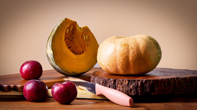 A studio still life composition of a sliced pumpkin, whole pumpkin, red plums, and a kitchen knife arranged on rustic wooden boards against a warm background. Ideal for concepts