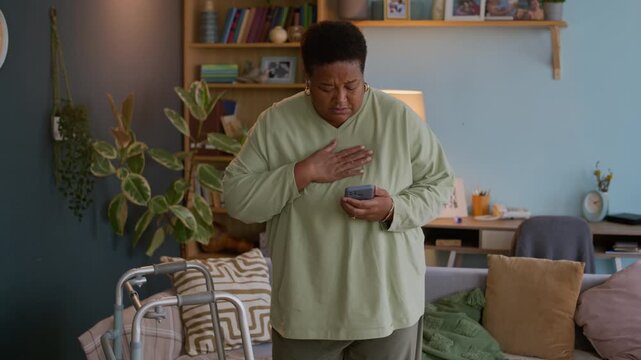 Medium long shot of African American elderly woman experiencing heart attack using smartphone while calling for help at home
