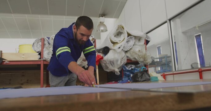 Leaning craftsman marking cutting lines on blue fabric at industrial table, with measuring tape