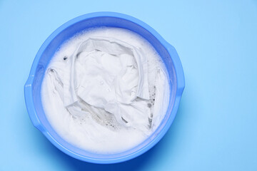 Plastic basin with clothes, water and foam on light blue background, top view