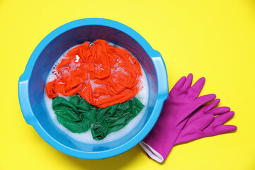 Plastic basin with clothes, water and gloves on yellow background, top view