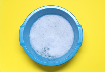 Blue plastic basin with water and foam on yellow background, top view