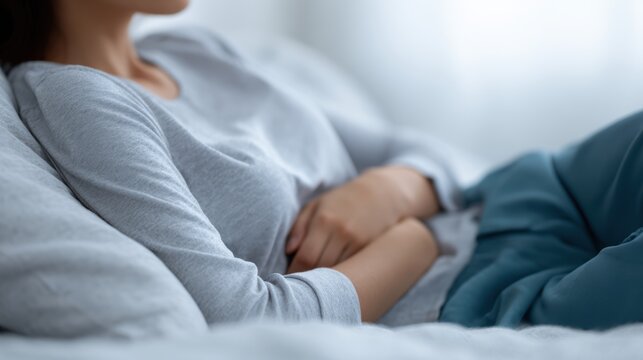 Sad young woman experiencing abdominal cramps during her period, lying on bed at home, holding her stomach. Menstrual discomfort and home care theme