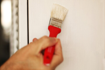 A man paints an old cabinet with a brush. He holds a red brush in his hand against the background of an old white cabinet.