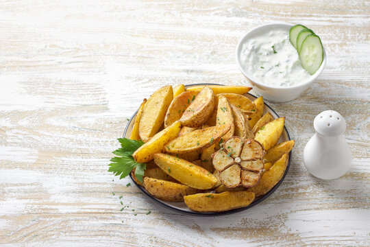 Golden baked potato wedges with garlic, spices and Greek tzatziki sauceon rustic plate. Tasty homemade snack or side dish, healthy vegetarian food