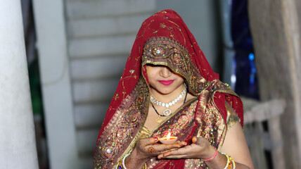 Beautiful Indian woman in traditional red saree holding a lit clay diya lamp, celebrating Diwali festival with prayerful expression in evening.