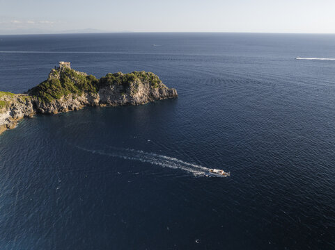 Aerial view of a boat slicing through the deep blue sea near a rocky outcrop topped with a historic tower, Conca dei Marini, Amalfi Coast, Italy.