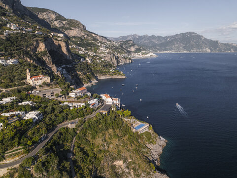 Aerial view of a dramatic coastline where the rugged cliffs meet the deep blue sea, dotted with white buildings and green foliage, Conca dei Marini, Amalfi Coast, Campania, Italy.