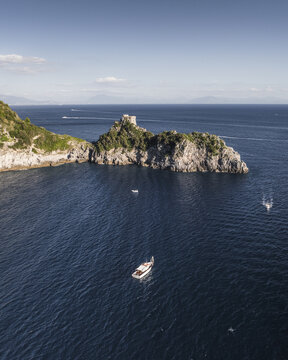 Aerial view of a rocky outcrop crowned with ancient fortifications, juxtaposed against the deep blue sea, where white yachts dance on the waves, Conca dei Marini, Campania, Italy.