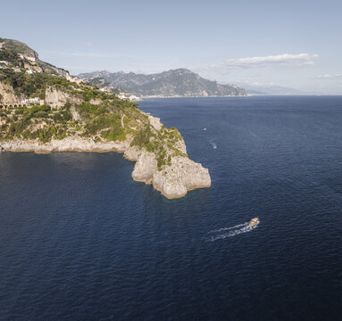 Aerial view of the rugged coastline with dramatic cliffs meeting the deep blue sea, a boat slicing through the water near the shore, Conca dei Marini, Amalfi Coast, Campania, Italy.