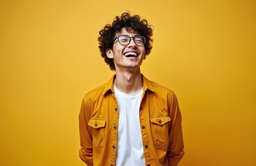 Fototapeta premium Joyful young man smiles widely. Male model wears glasses and yellow shirt. Happy guy with curly hair laughs against yellow backdrop. Positive person looks at camera.