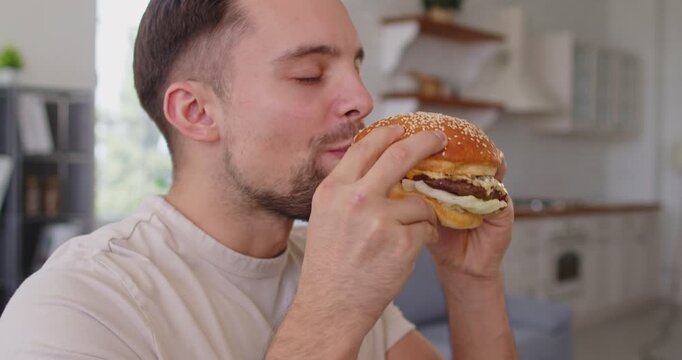 Hungry man eating burger, happy young person holding sandwich with fresh buns and meat to bite with closed eyes and taste, enjoy delicious fast food snack. Satisfaction of guy with big hamburger