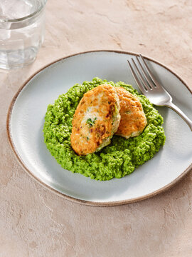 Golden chicken patties are placed atop a smooth layer of creamy smashed peas, creating an appetizing dish on a clean plate. A refreshing glass of water is nearby