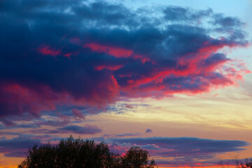 A high-detail shot of the sky showcases dramatic clouds with deep blue and crimson hues, highlighted by a vibrant pink and red glow. Softer layers of yellow and orange are visible below, with a silhou