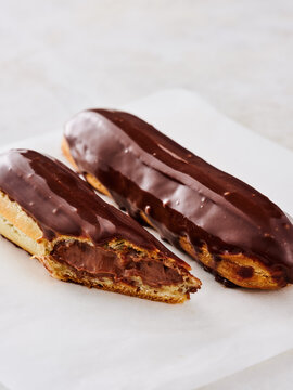Two freshly made chocolate eclairs sit on a clean white surface, one cut open to reveal its rich, creamy filling. Perfect for dessert lovers at any time of day