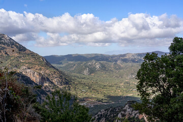 Naklejka premium Beautiful panoramic view of the Cretan countryside with mountains, green valleys, olive groves, and a small traditional village under a bright blue sky with scattered clouds. Captured on a sunny day