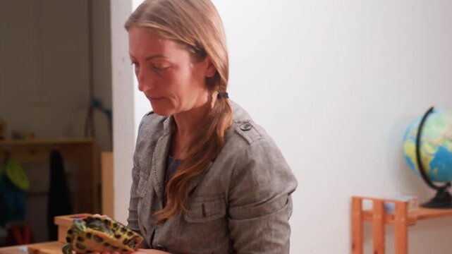 Woman in gray jacket holding tortoise carefully while interacting with someone in indoor setting, preparing to bend down and place animal, educational moment