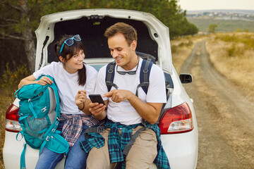 Couple by car use phone, map, backpack. Stranded on a rural road, travelers check geolocation, seek signal, and study a map while resting on trunk near forest. Adventure on weekend travel and hiking.