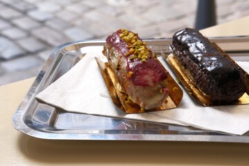 Two eclairs on metal tray with napkins on table of oudoor cafe, blurred street on background. Raspberry and chocolate classic french dessert. Soft focus