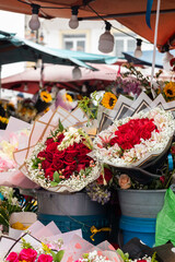 Beautiful flower bouquets with red roses, white blooms, and sunflowers arranged in buckets at an outdoor flower market. The vibrant colors and lively atmosphere capture the charm of a local floral mar