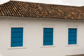 A serene colonial-style wall with vibrant blue windows stands beneath a weathered clay tile roof, evoking cultural heritage, simplicity, and timeless architectural charm.
