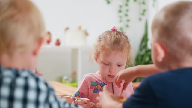 Close up rear view of boy in blue pulling paint container toward himself during art class as girl in pink shirt with flower design attentively watches, children concentrating on creative activity