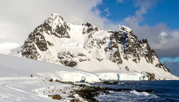 A snow-covered mountain range with a rugged peak towers over a frozen landscape, meeting the icy ocean under a cloudy sky
