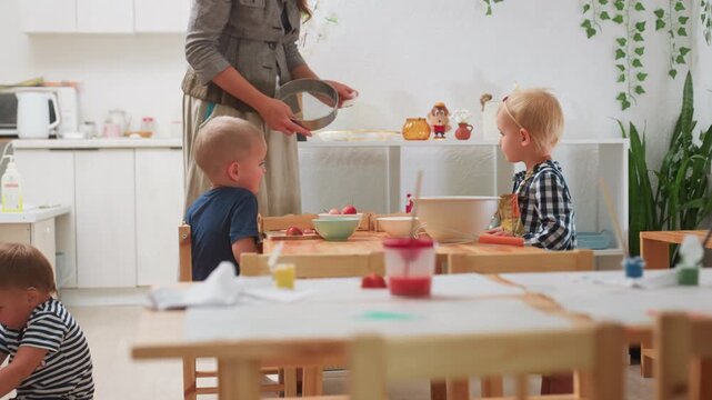 Mother brings sift while collecting chopper from child during kitchen activity as children sit at wooden table with bowls and ingredients, creating moment of guidance, learning, and shared experience