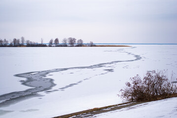 Frozen lake landscape with melting ice and bare trees in winter season