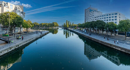 Obraz premium Paris, France - 10 11 2025: Ourcq Canal. Panoramic view of reflections on the Ourcq canal of trees, bridge and buildings at sunrise